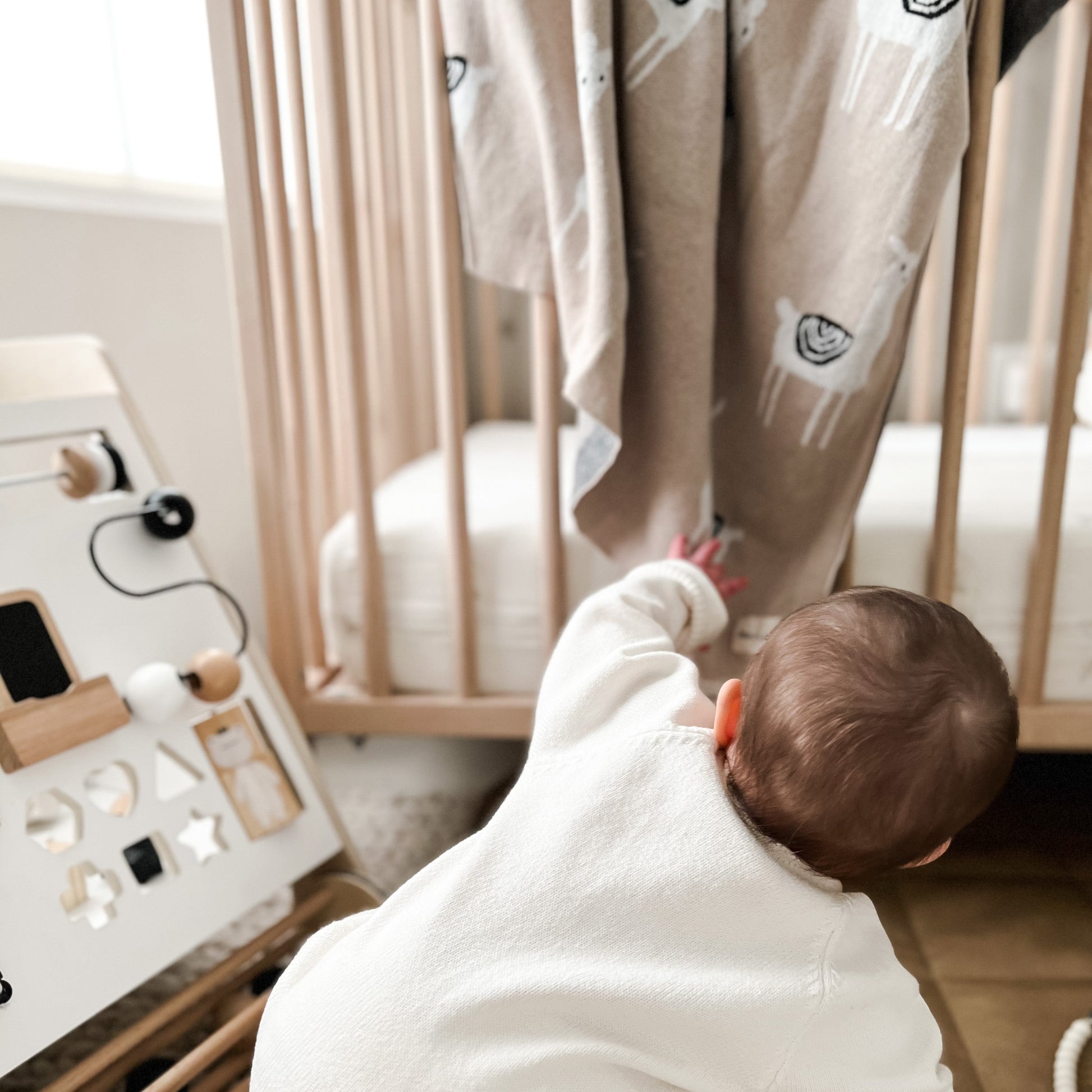 A baby in a white outfit is reaching towards a wooden toy with llama shapes.