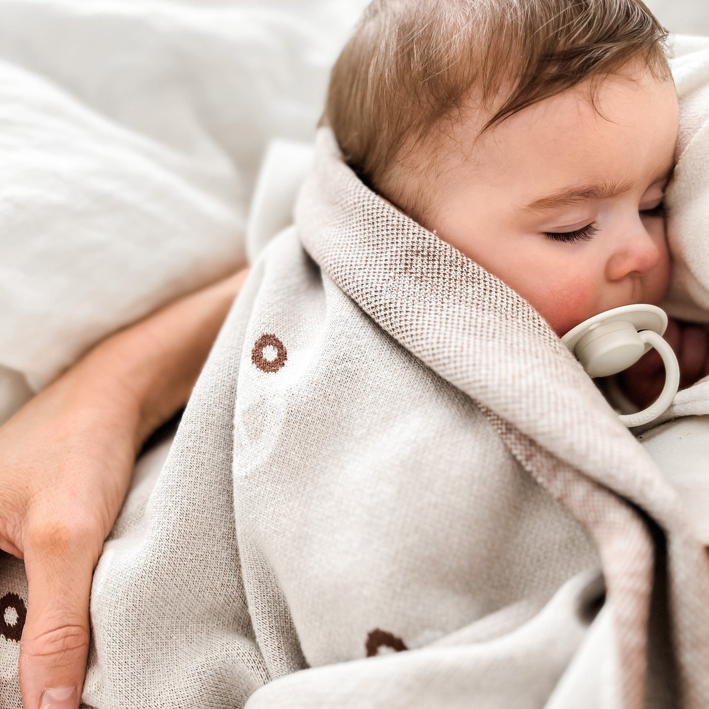 Baby wrapped in a beige blanket with a pacifier, held by an adult.