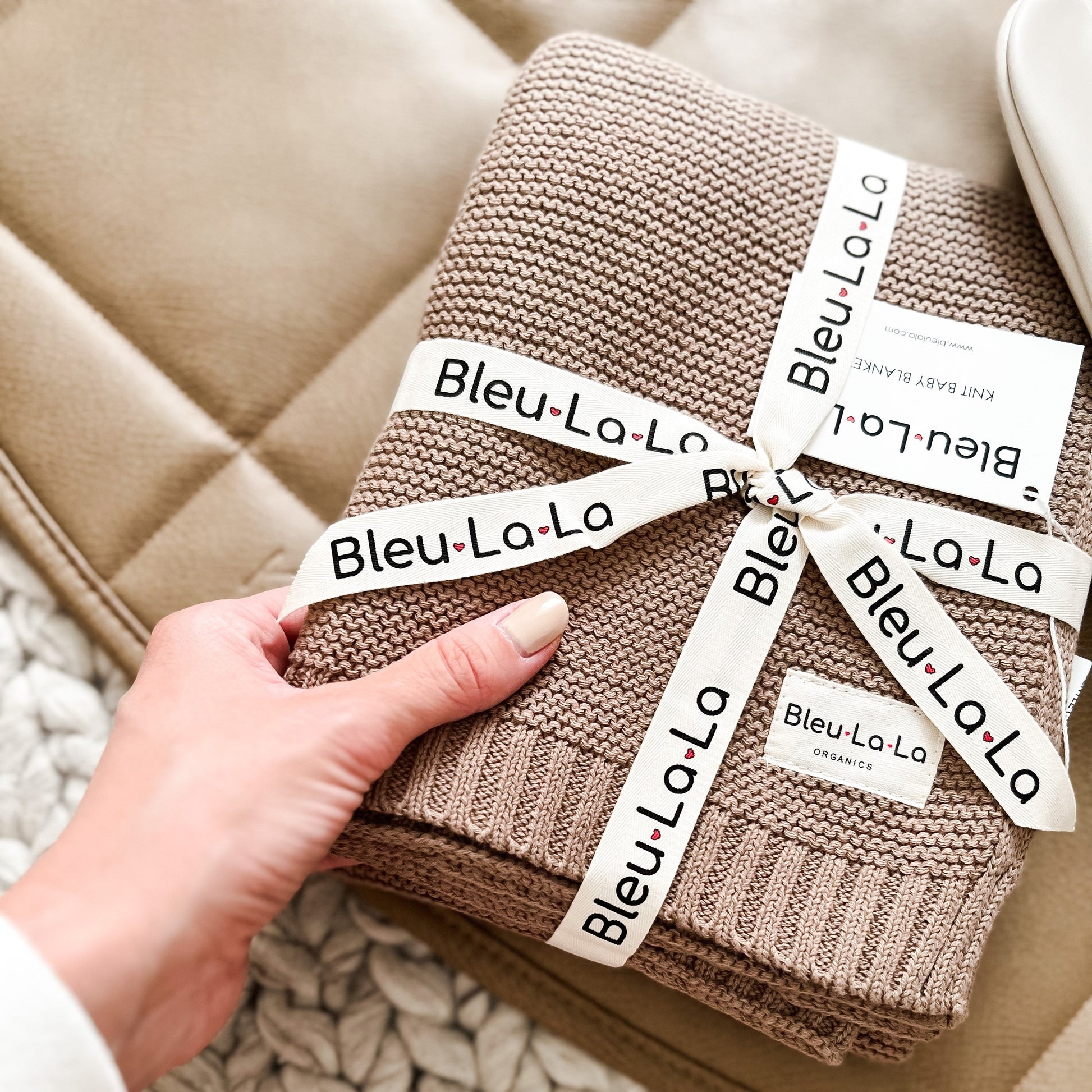 Beige knitted blanket with 'Bleu La La' labels held by a hand against a beige sofa.