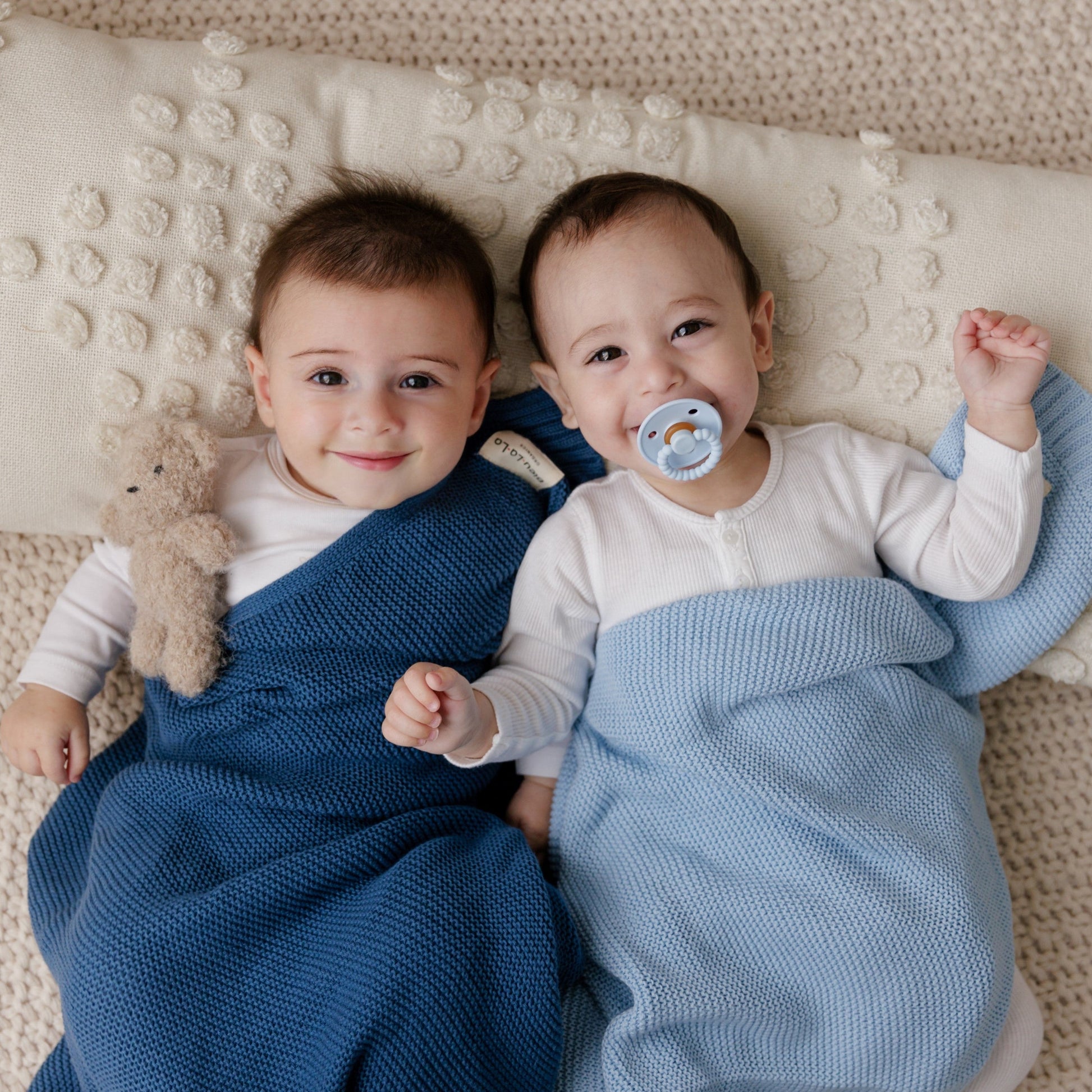 Two babies lying on a textured blanket, one with a blue  Blue-La-La blanket and the other with a blue pacifier.