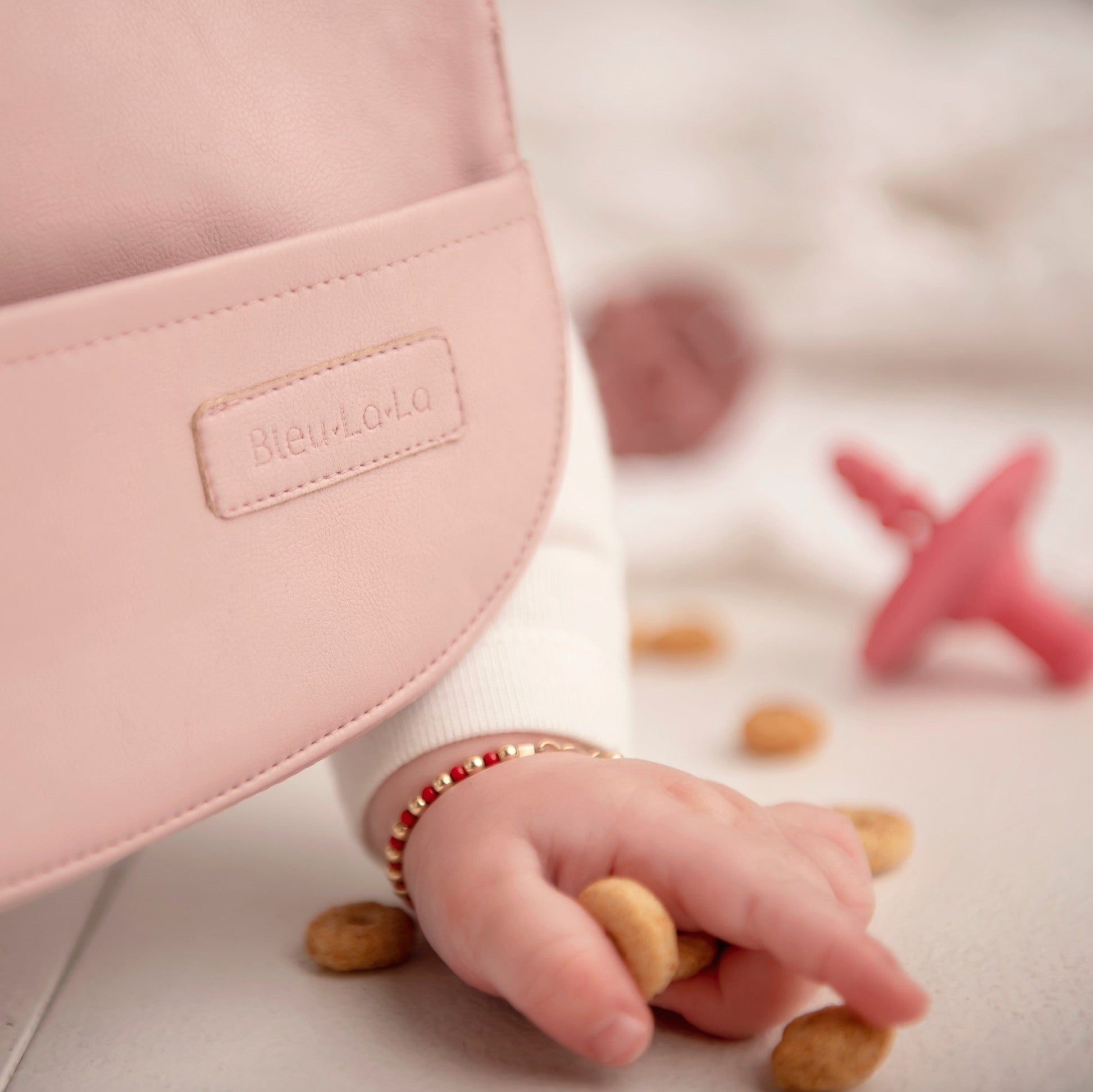 Close-up of a baby's hand holding a cookie near a pink bag with 'BleuLola' branding.