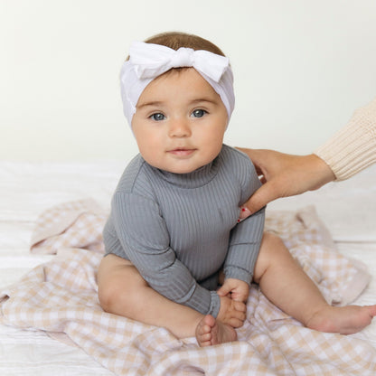 Baby wearing a gray ribbed outfit and white headband with a bow, sitting on a checkered blanket.