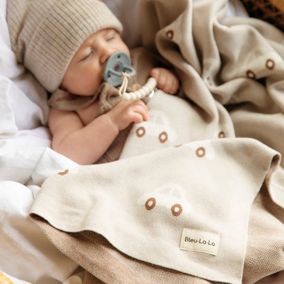 Baby wrapped in a blanket with a 'Bleu-La-La' label, wearing a beige knit hat and holding a blue pacifier.