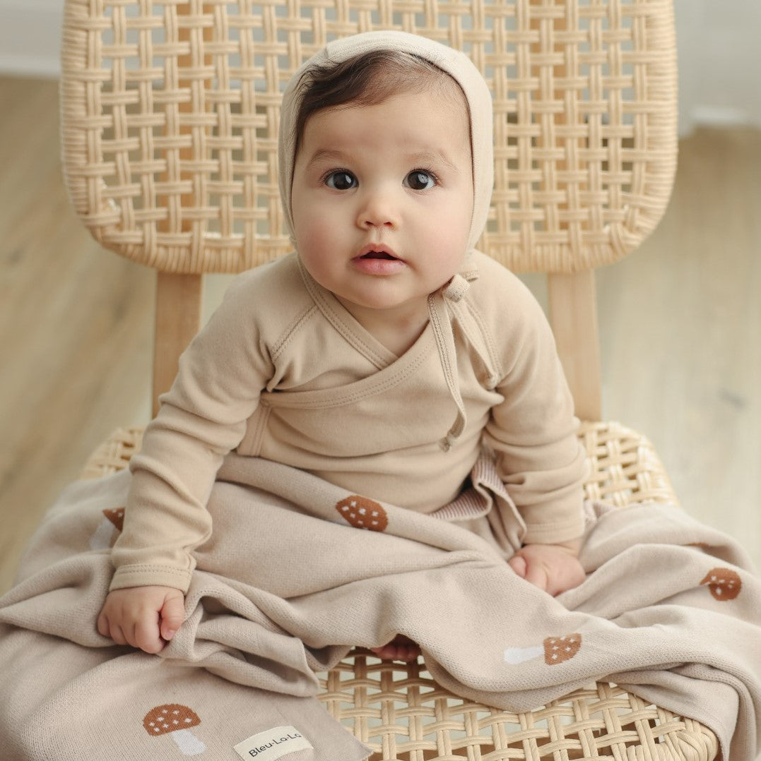 A baby in a beige outfit with a hoodie and luxury knit mushroom blankets is sitting on a wicker chair.
