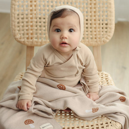 A baby in a beige outfit with a hoodie and luxury knit mushroom blankets is sitting on a wicker chair.