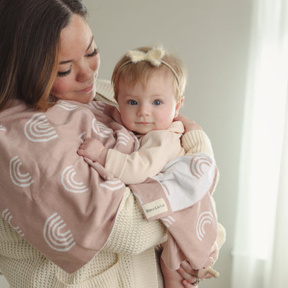 Woman holding a baby wrapped in a pink blanket with white rainbow patterns, indoors.