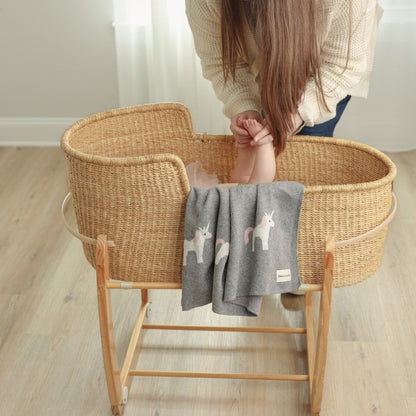 Person holding a child's hand inside a wicker crib with a gray blanket featuring unicorn designs.