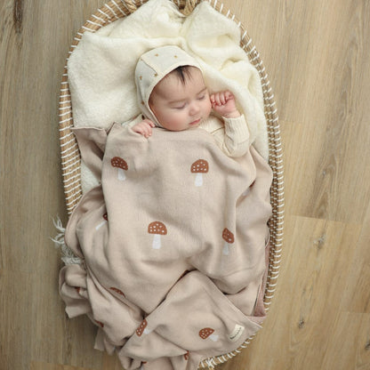 Baby wrapped in a beige blanket with mushroom patterns, lying in a wicker basket on a wooden floor.