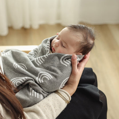 Newborn baby wrapped in a gray blanket with white rainbow patterns, held by an adult in a softly blurred indoor setting.