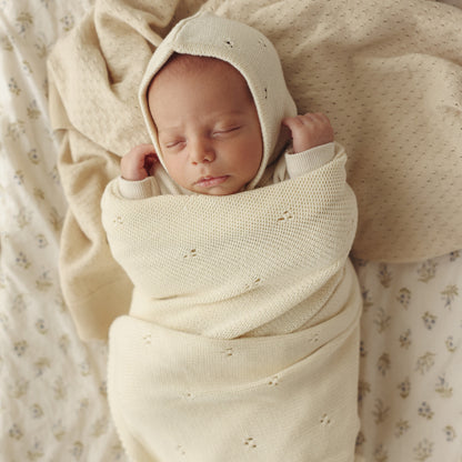 Newborn baby wrapped in a cream-colored swaddle and bonnet on a soft, patterned blanket.