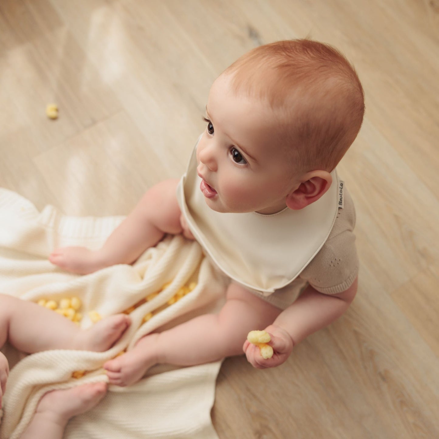 Baby sitting on a wooden floor with a white blanket wearing a soft vegan leather easy clean baby bibs with adjustable snaps