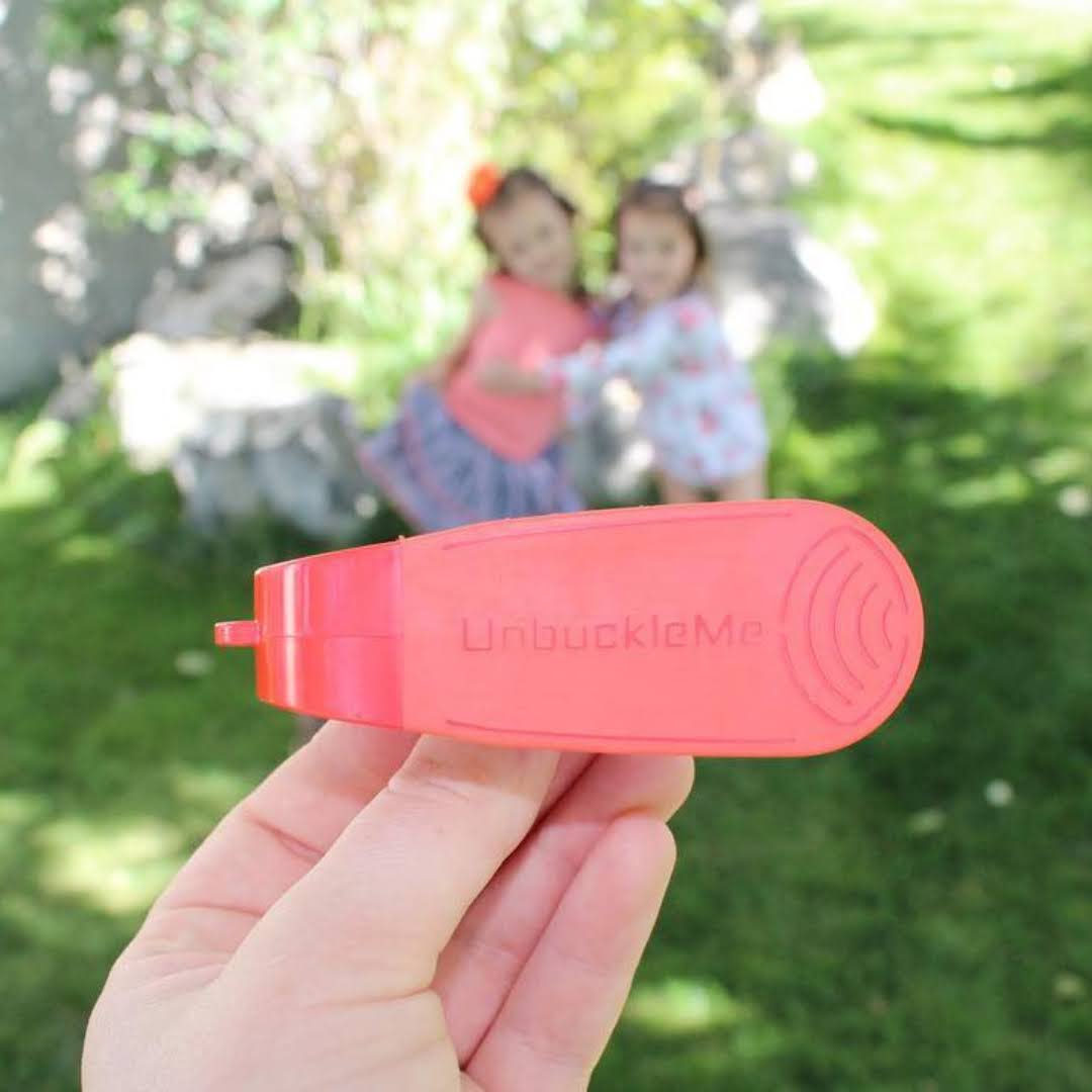 Red 'UnbuckleMe' device held in a hand with children playing in the background.