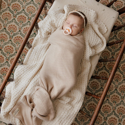 Newborn baby swaddled in beige blanket with pacifier, lying in a wooden crib.