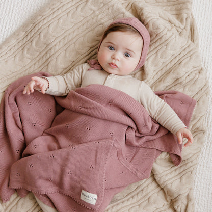 Baby lying on a textured surface with a pink blanket and headband, wearing a beige outfit.
