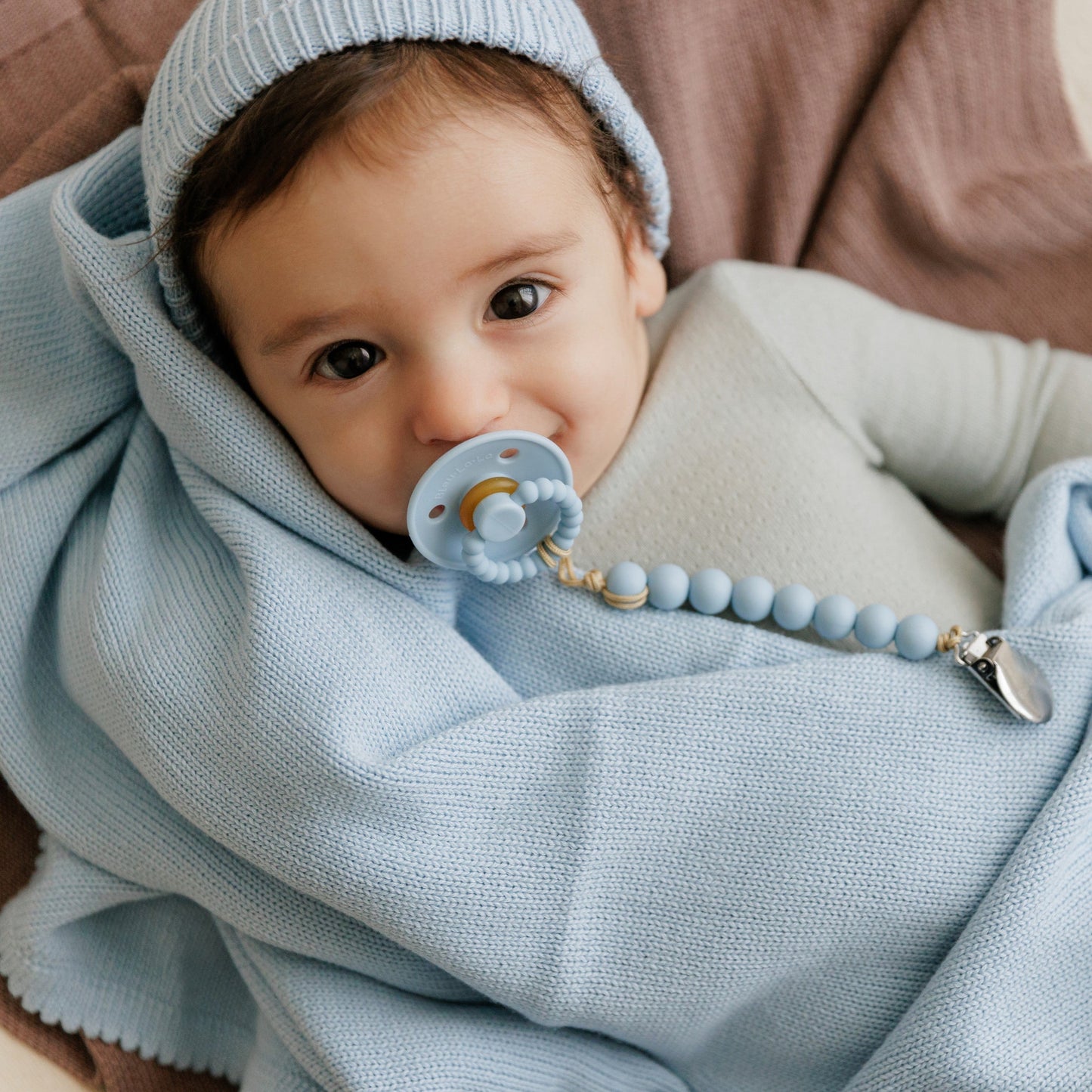Baby wrapped in a light blue blanket with a blue Luladream premium pacifier, wearing a matching hat.
