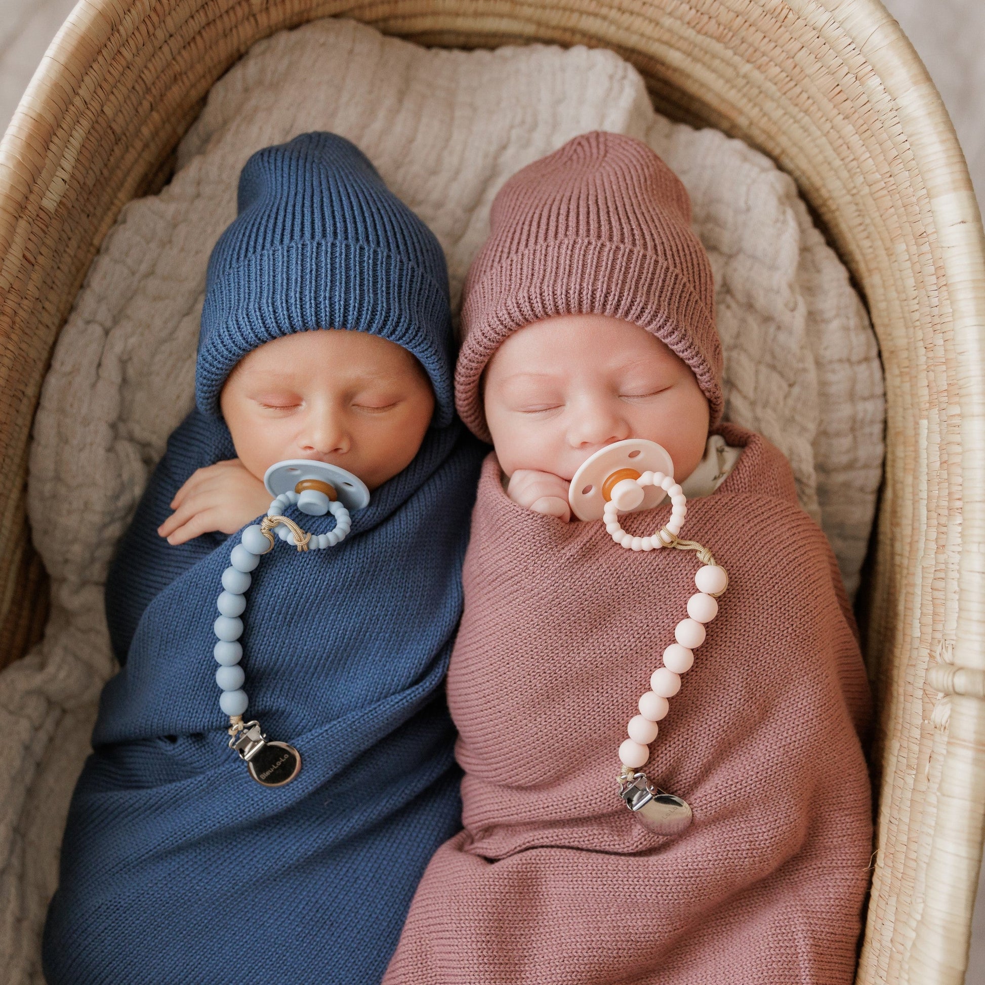 Two babies swaddled in blue and pink  each with Luladream premium pacifier, wearing knit hats.