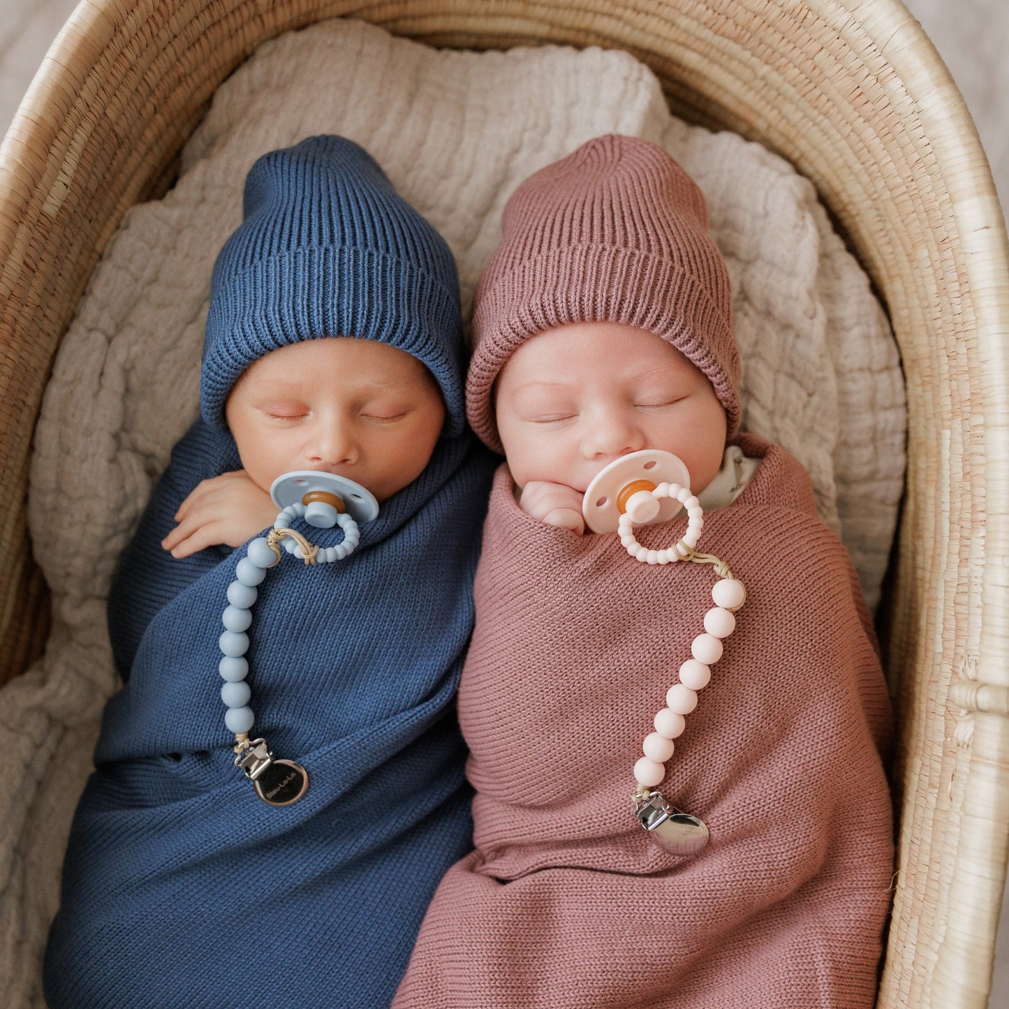 Two babies swaddled in blue and pink with pacifiers, wearing knit hats.