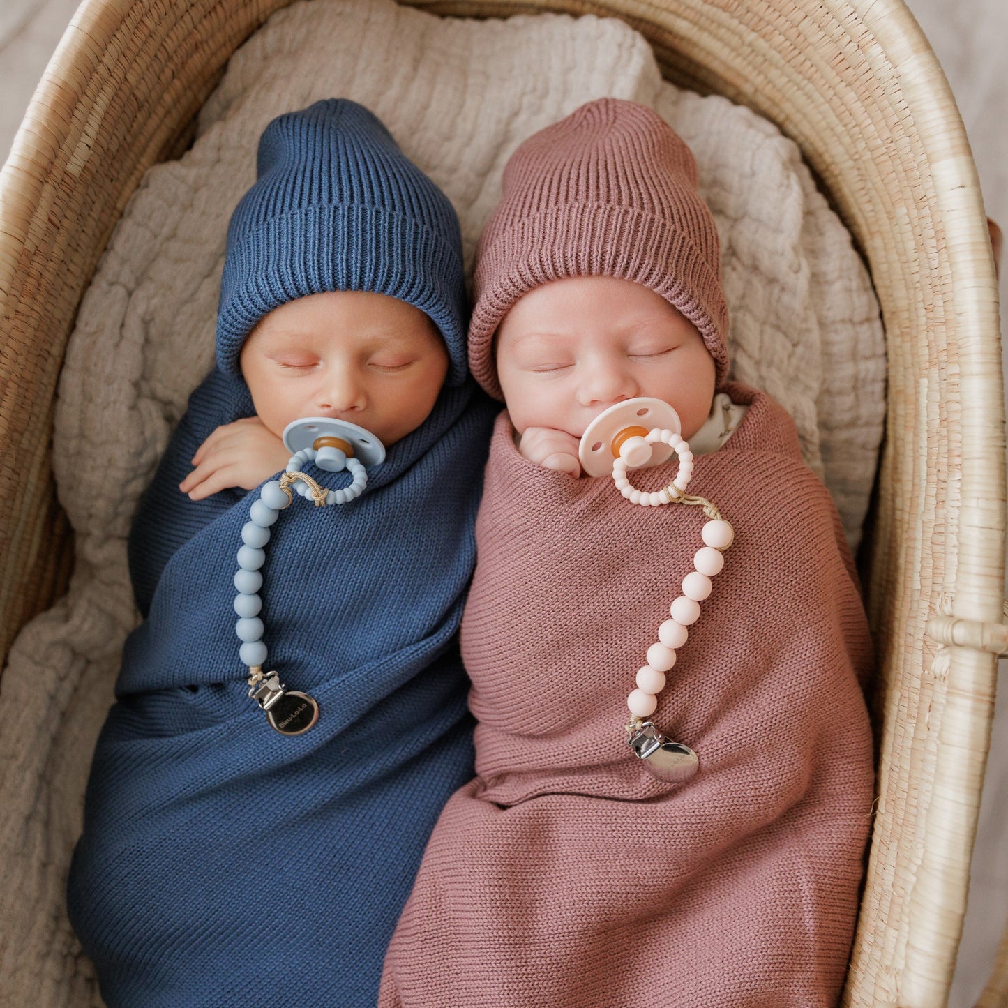 Two swaddled babies in Bleu-la-la blankets wearing knit hats and pacifiers in a basket.