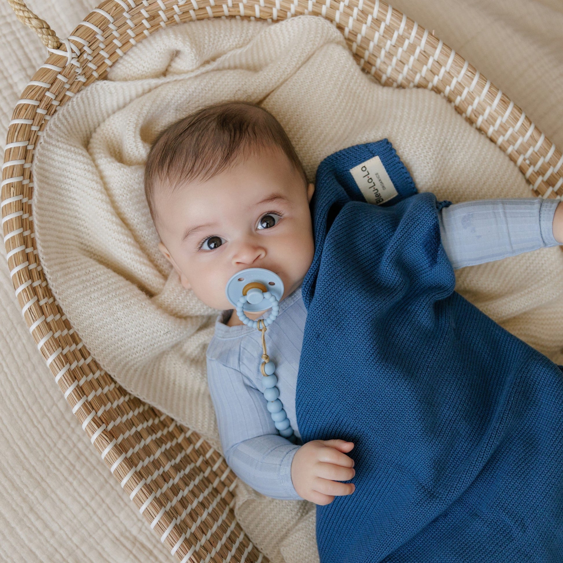 Baby wrapped in a blue blanket with a Luladream Pacifier pacifier, lying in a woven basket.