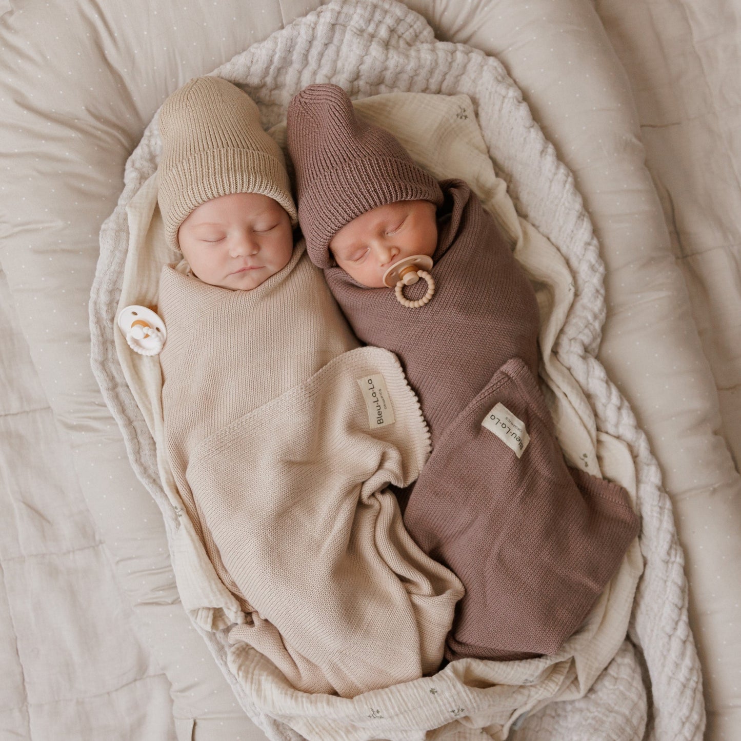 Two newborn babies wrapped in beige and brown swaddles with hats and pacifiers, lying on a soft surface.