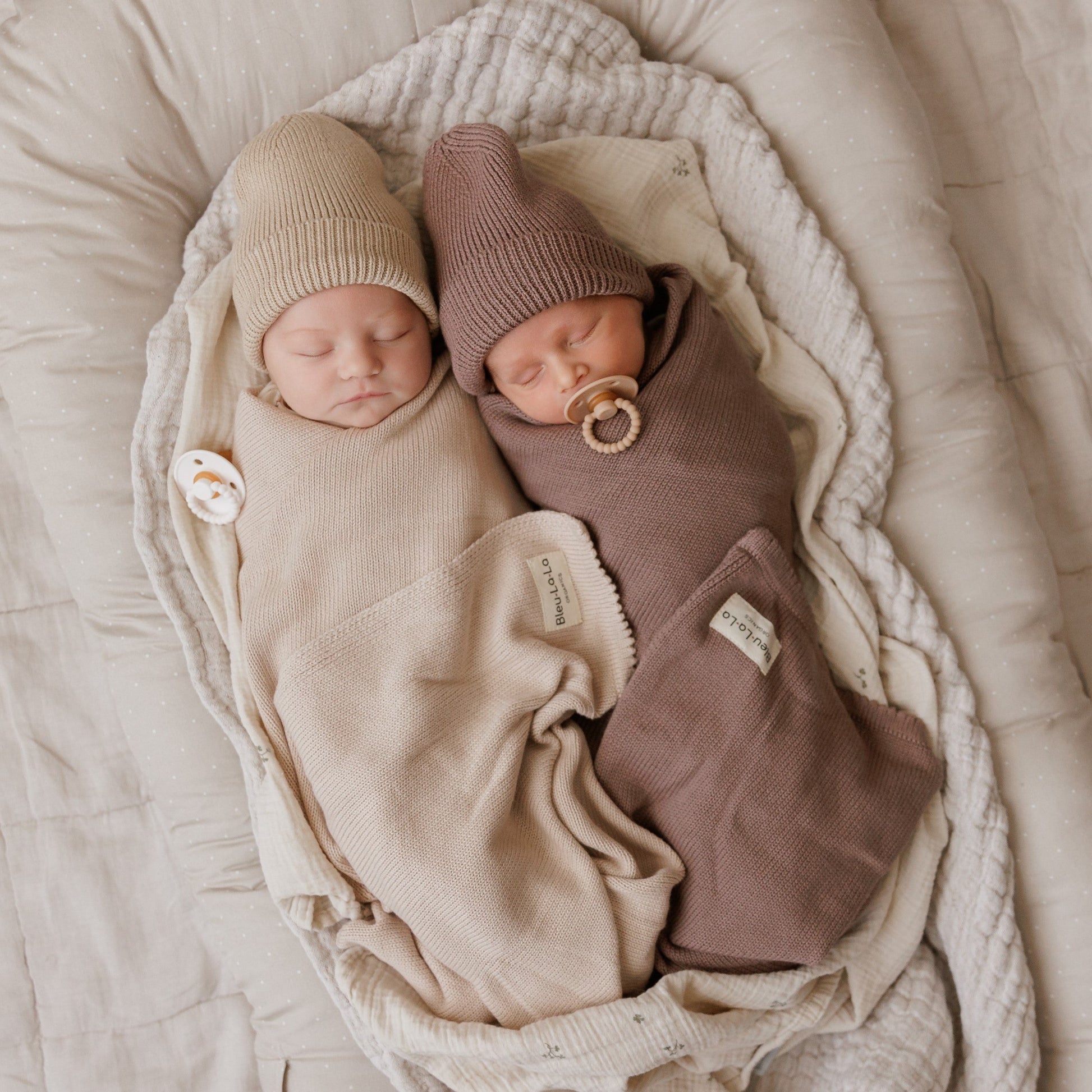 Two newborn babies wrapped in beige and brown swaddles with hats and pacifiers, lying on a soft surface.