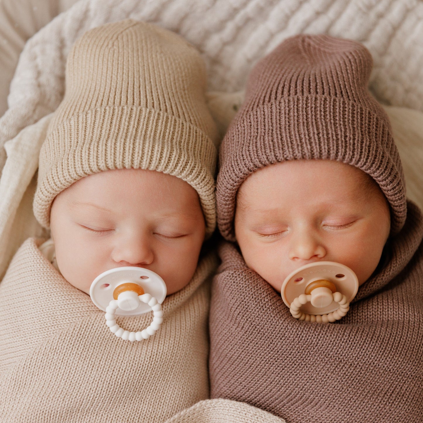 Two newborn babies wearing knit hats and using Luladream premium pacifiers lying next to each other on a soft surface.