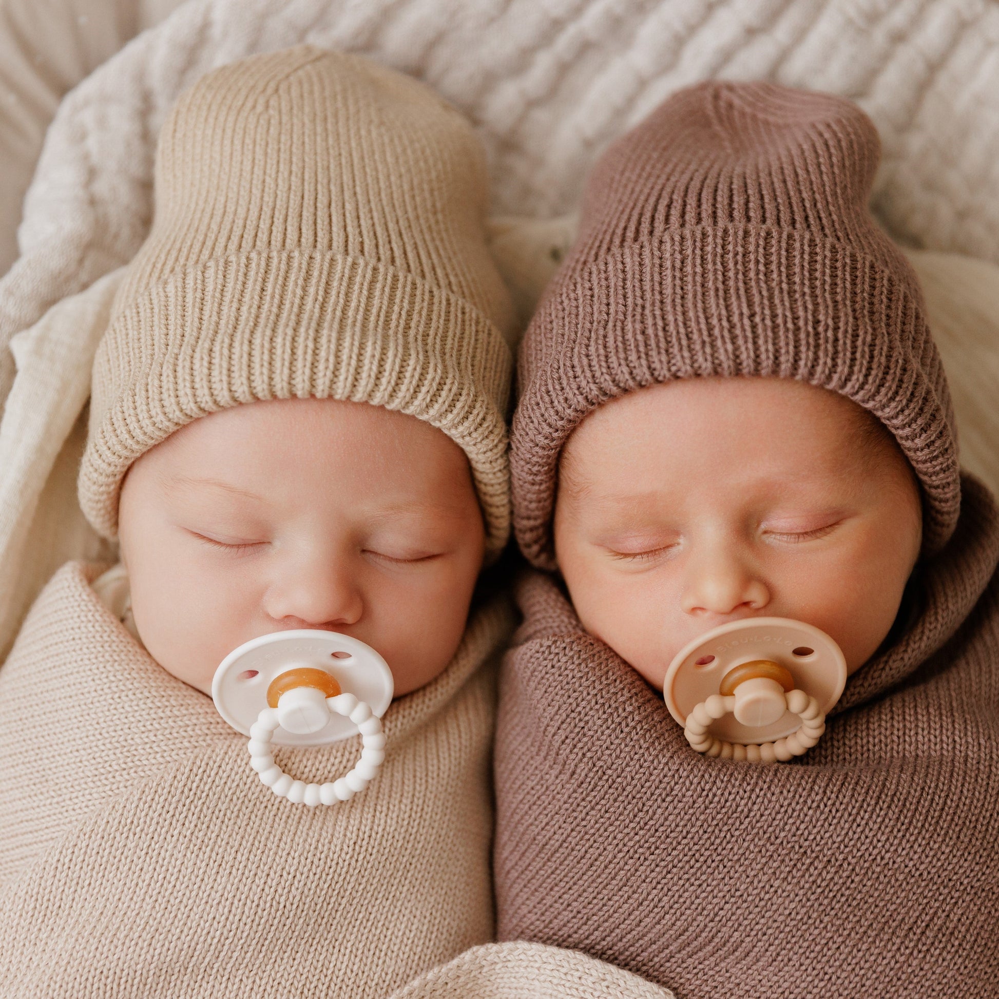 Two newborn babies wearing knit hats and using Luladream premium pacifiers lying next to each other on a soft surface.