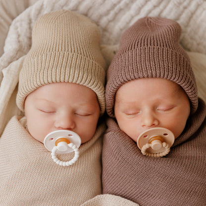 Two newborn babies wearing knit hats and using Luladream premium pacifiers lying next to each other on a soft surface.