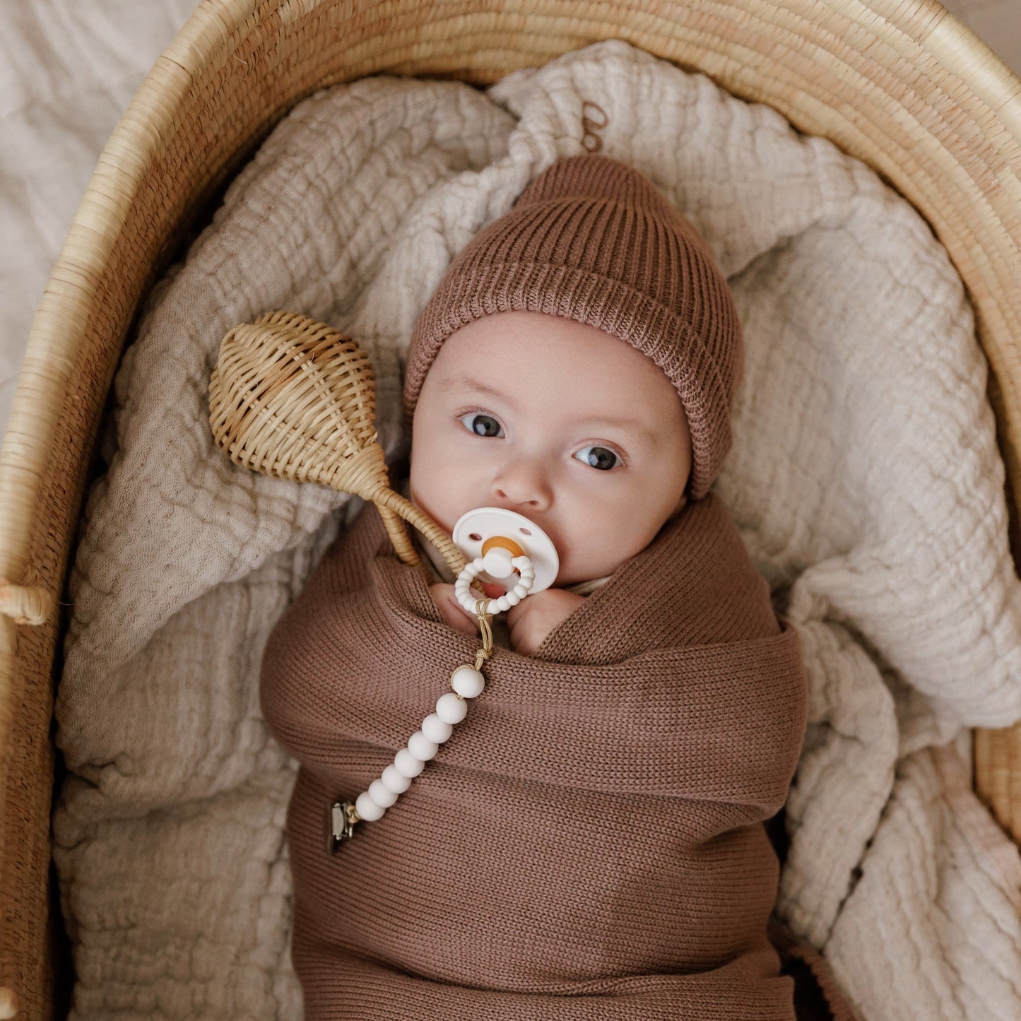 Baby wrapped in brown fabric with a Luladream pacifier lying in a woven basket.