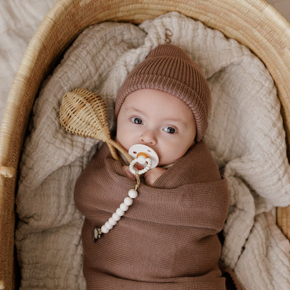 Baby wrapped in brown blanket with a Luladream Pacifier pacifier, lying in a woven basket.