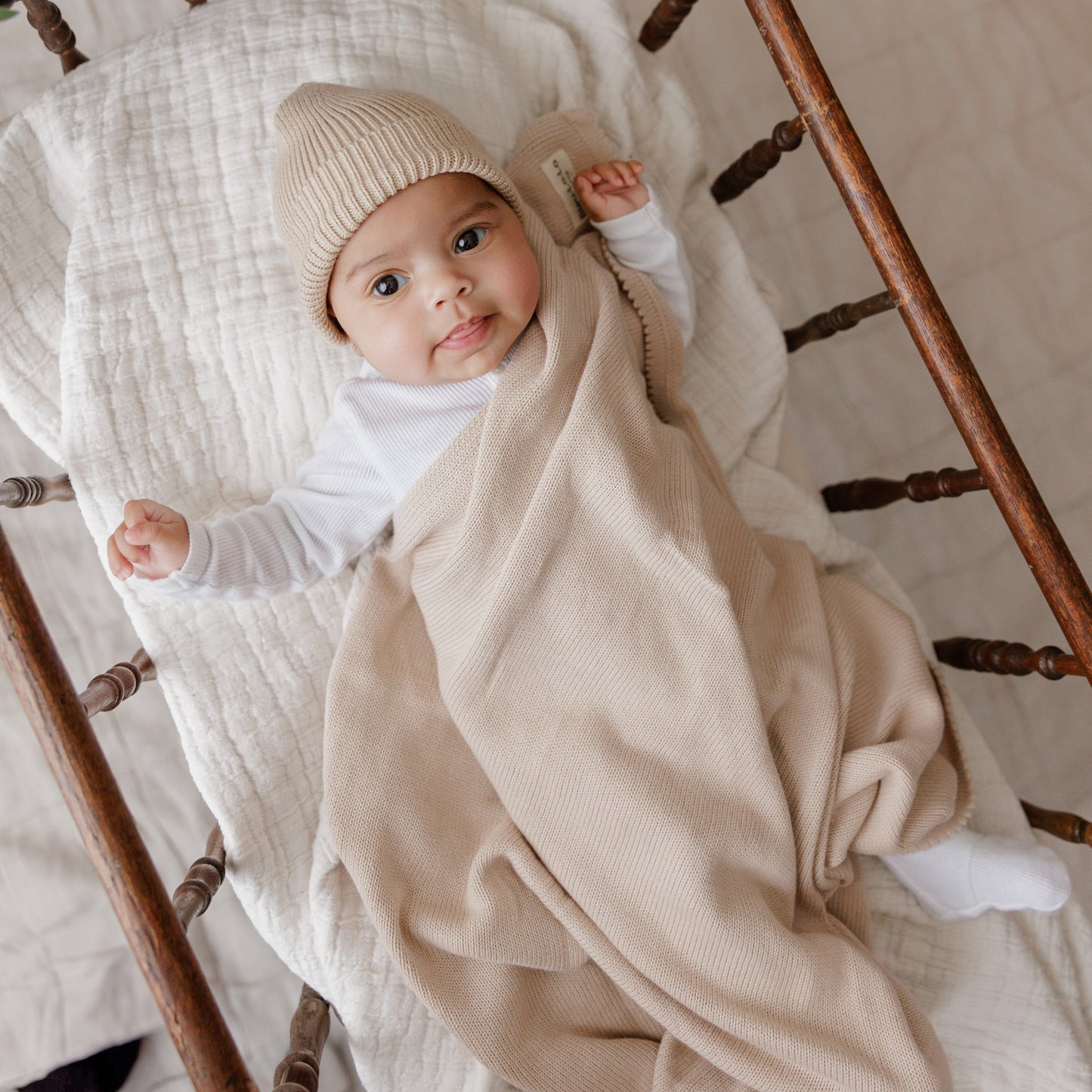 Baby wrapped in a beige blanket and hat, sitting on a wooden chair.