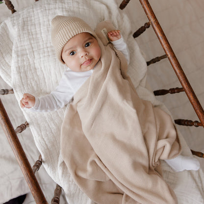 Baby wrapped in a beige blanket and hat, sitting on a wooden chair.