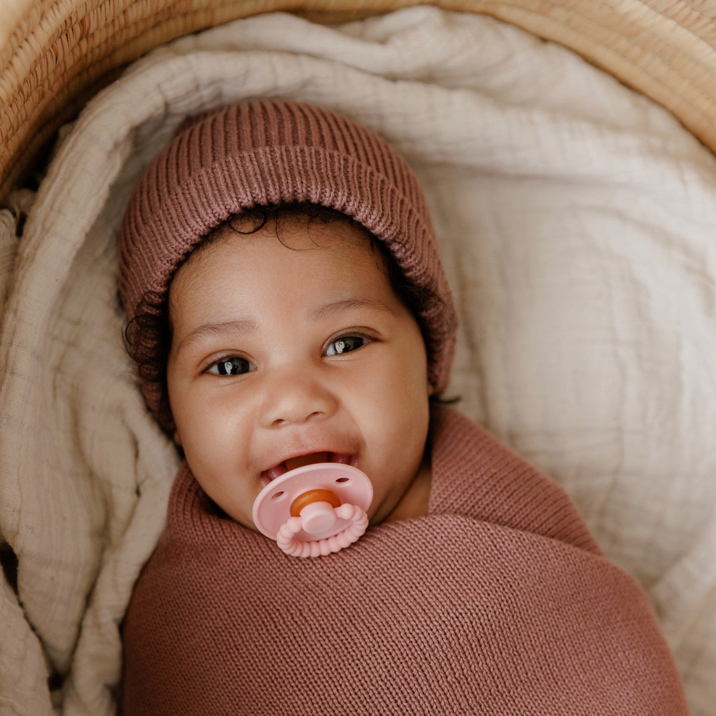 Baby wrapped in pink blanket with pink Luladream pacifier and hat, lying on beige fabric