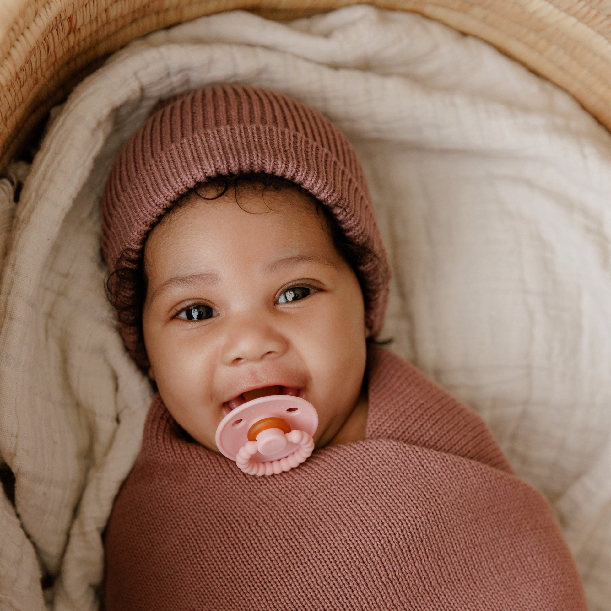 Baby wrapped in pink blanket with pink pacifier and hat, lying on beige fabric