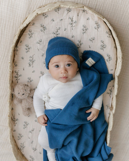 Baby in a floral Moses basket wearing a blue hat and blanket Bleu-la-la