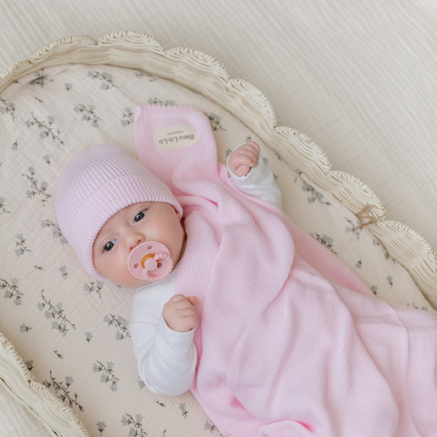 A newborn baby in a pink outfit and hat is lying in a crib with a pacifier. Bleu-la-la.