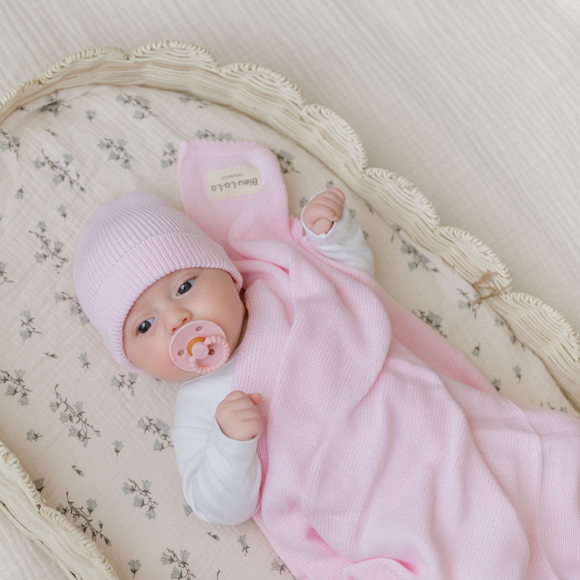 A newborn baby in a pink outfit and hat is lying in a crib with a pacifier. Bleu-la-la.