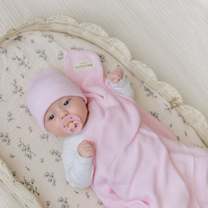 A newborn baby in a pink outfit and hat is lying in a crib with a pacifier. Bleu-la-la.