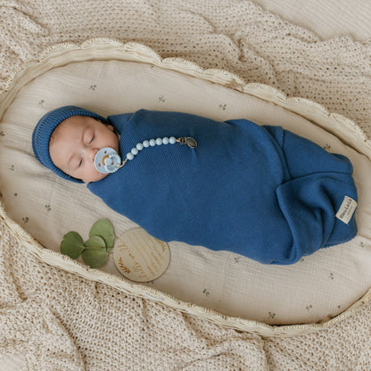 Newborn baby wrapped in blue swaddle with pacifier and hat, lying on a textured surface.
