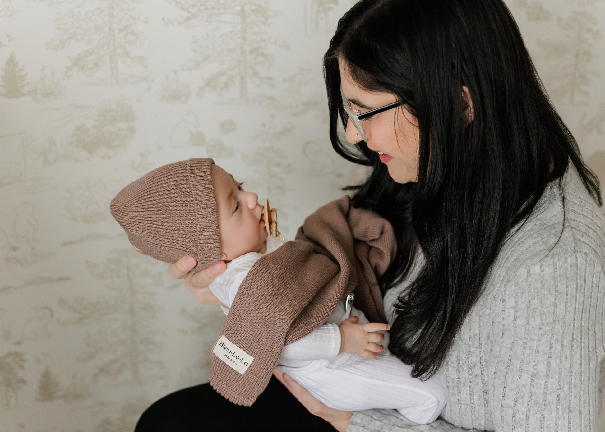 Woman holding a baby wrapped in a brown blanket from Bleu-la-la, with a neutral background