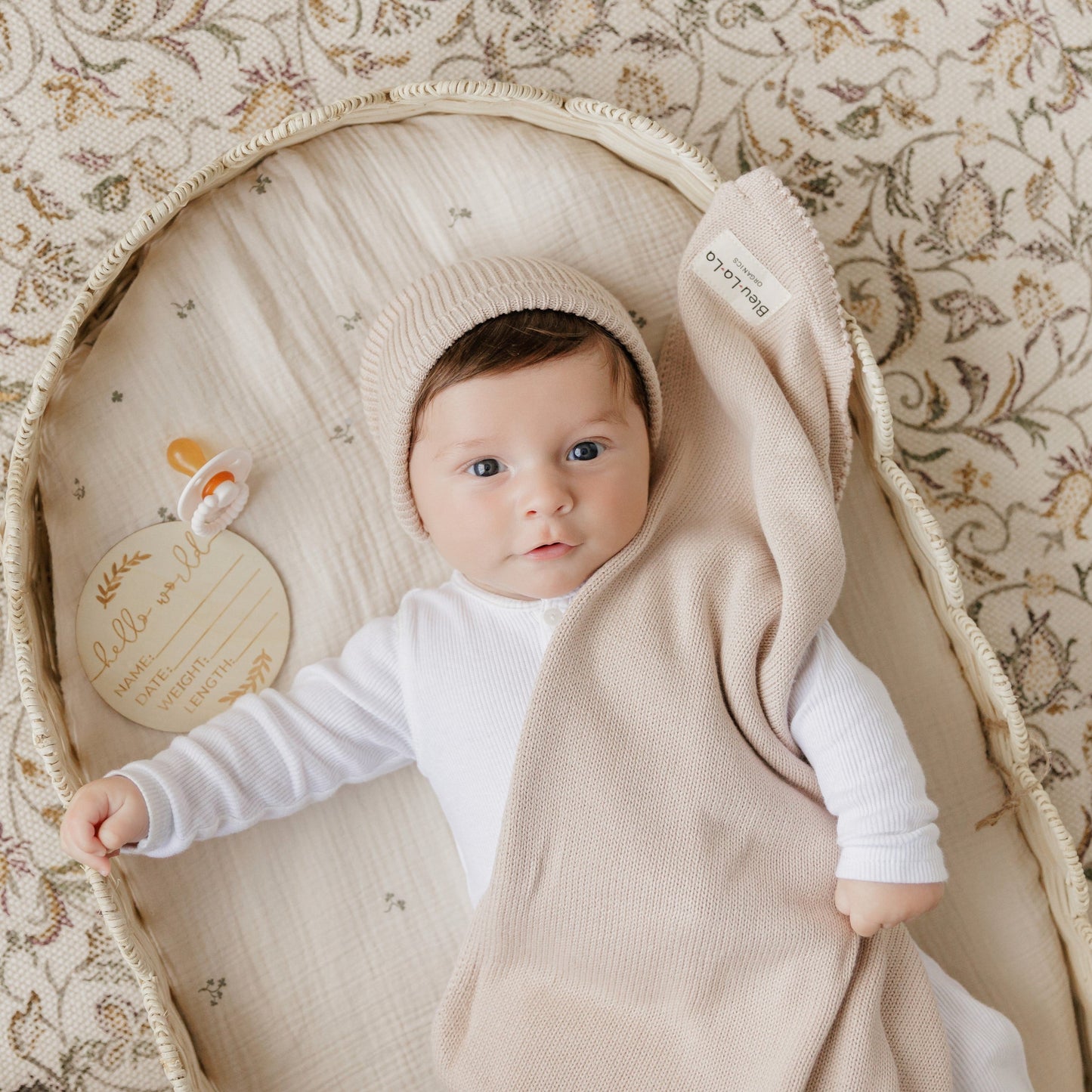 Baby in a beige swaddle and hat lying in a circular crib with a pacifier on a floral-patterned blanket.