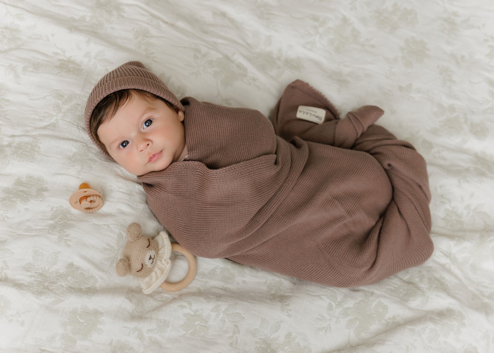 Newborn baby wrapped in a brown swaddle and hat from Bleu-la-la, lying on a white blanket with toys nearby.