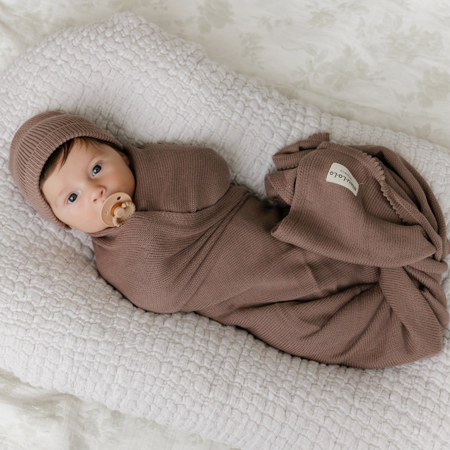 Newborn baby wrapped in a brown swaddle with matching hat on a textured white blanket from Bleu-la-la.