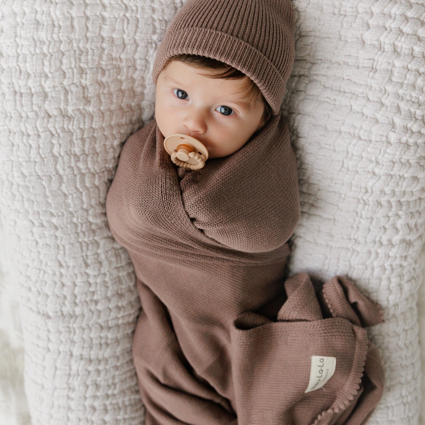 Newborn baby wrapped in a brown swaddle and hat from Bleu-la-la, lying on a textured white blanket.