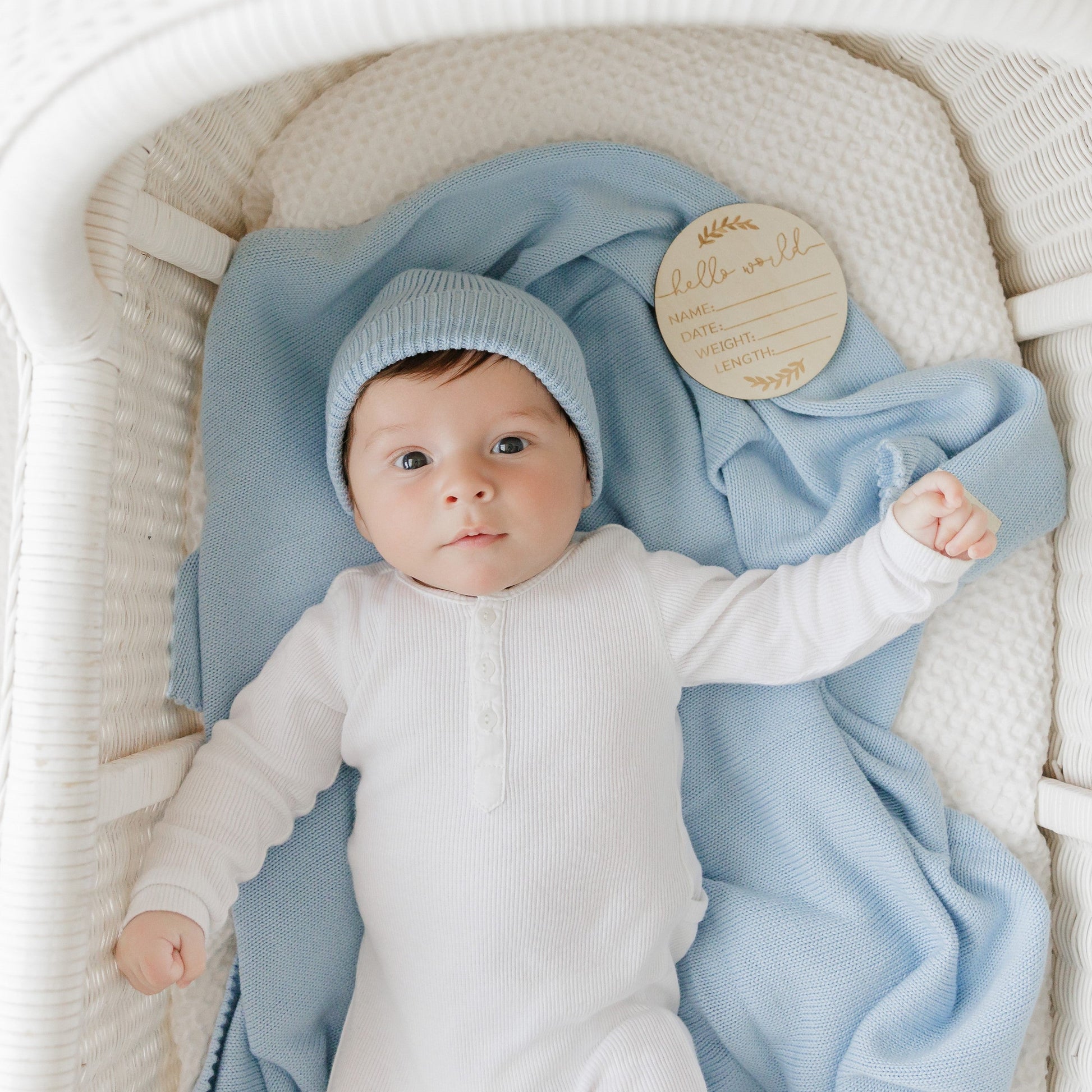 Baby in a white outfit and blue hat lying on a blue blanket from Bleu-la-la, in a white crib.
