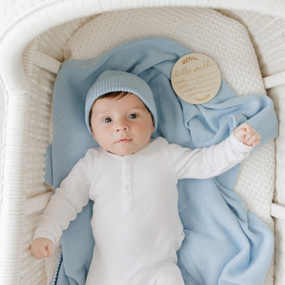 Baby in a white outfit and blue hat lying on a blue blanket from Bleu-la-la, in a white crib.