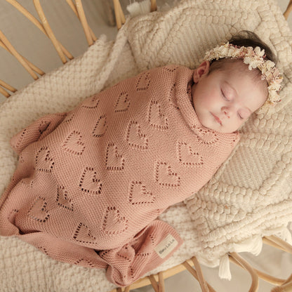 Newborn baby wrapped in a pink Bleu-la-la knitted blanket with heart patterns, lying on a textured surface.