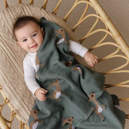 Child wrapped in a green blanket with deer patterns, sitting in a wicker chair.