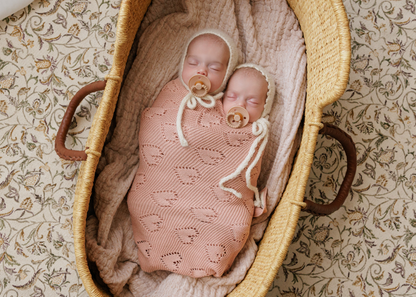 Two babies wrapped in pink  Bleu-la-la blankets with pacifiers in a wicker basket on a patterned surface.