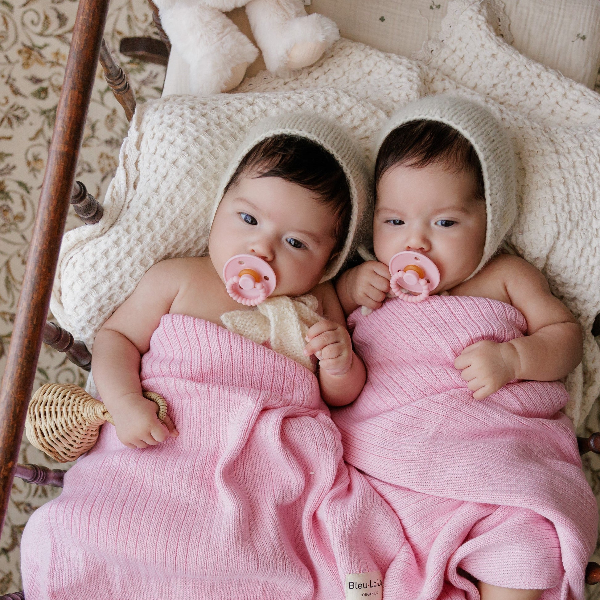 Two babies wrapped in pink blankets with Luladream premium pacifier set, wearing bonnets, in a cozy setting.